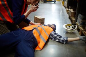 Male warehouse staff in high visibility vests. One worker is unconscious on the floor and the other is radioing for help.