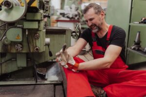 A worker in red overalls sat on the floor next to a machine holding their hand in pain