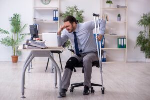 A man in business wear sits at a desk in an office. He has a broken leg and crutches.