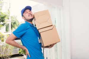 A man carrying some boxes and holding his lower back in pain due to suffering a manual handling injury.