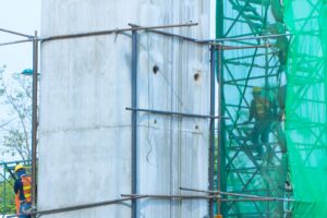 Scaffolding surrounding a large concrete pillar with green netting on the scaffolding for employee safety. 