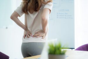 A woman with a back ache standing with her hands on her back in an office. 