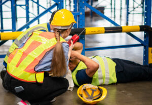 A foreman tends to a colleague lying on the floor in a warehouse who has had an accident at work.