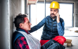 An injured worker with blood on their head and arm in a sling while a coworker is on the phone